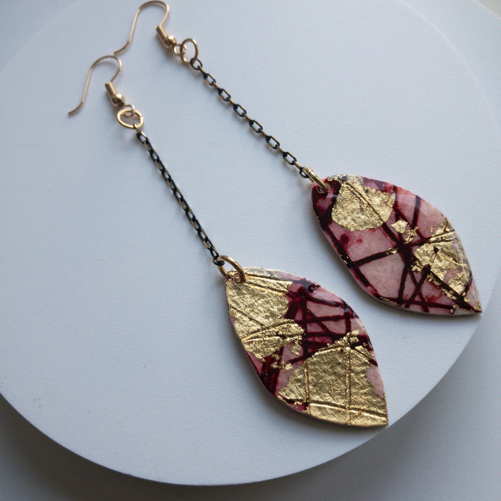 The Rothlú Gile Abstract Leaf Batik Textile Earrings in Red/Gold feature marquise-shaped pendants with a unique marbled pink texture, gold leaf patterns, and dark red abstract lines. These Sgraffito batik earrings are attached to hypo-allergenic gold-plated hooks by thin black chains and are displayed on a circular white surface.