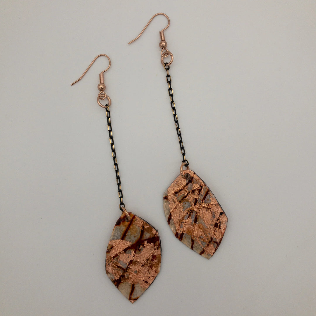 Rothlú's Gile Abstract Leaf Batik Textile Earrings in Rust/Rose Gold feature unique handmade leaf-shaped pendants with a marbled pattern, hanging from short black chains and copper hooks, shown on a white background.