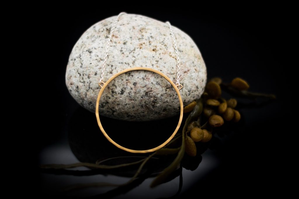 A delicate Ancaire Yellow Gold Vermeil Neckpiece by Lynsey de Burca displayed against a smooth, speckled stone. The thin chain and minimalist design of the unique jewelry create a simple yet elegant look. The stone serves as a natural, contrasting backdrop, with small twigs and buds positioned nearby.