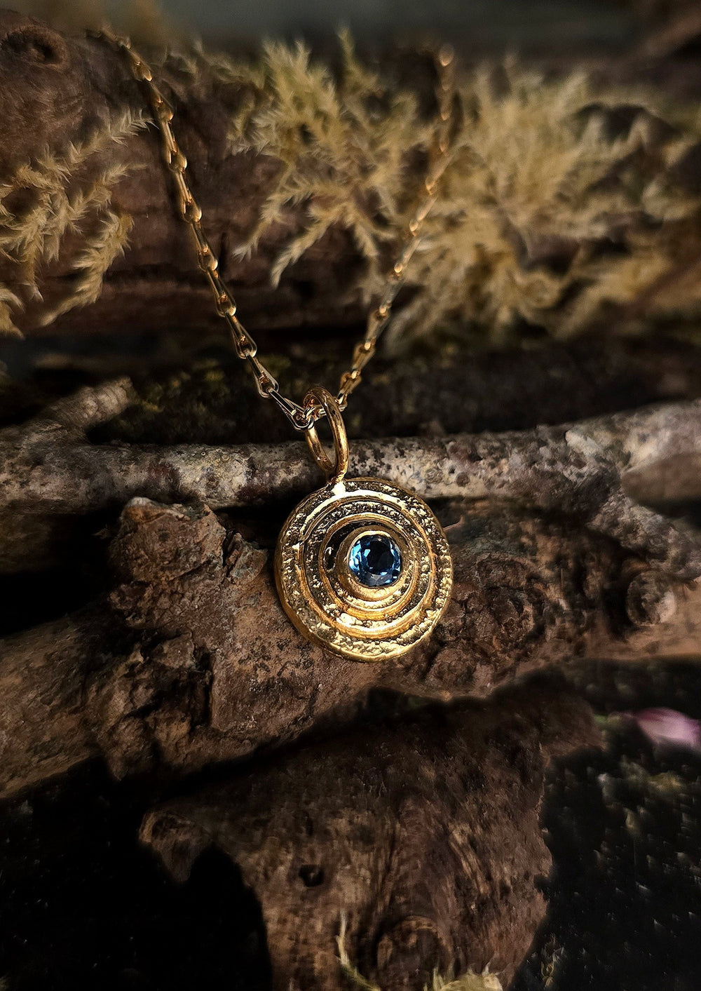 The Orb Pendant Gold in Blue Topaz by Caroline Stokesberry-lee features concentric circular designs and a blue topaz center, shown on textured wood and moss in natural, earthy lighting.