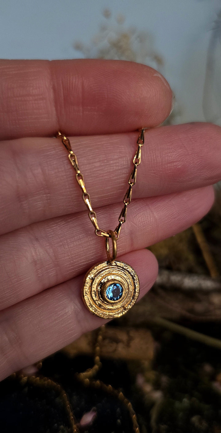 A hand holds the Caroline Stokesberry-lee Orb Pendant Gold in Blue Topaz—a gold-plated sterling silver necklace featuring a central blue topaz gemstone—against a blurred background.