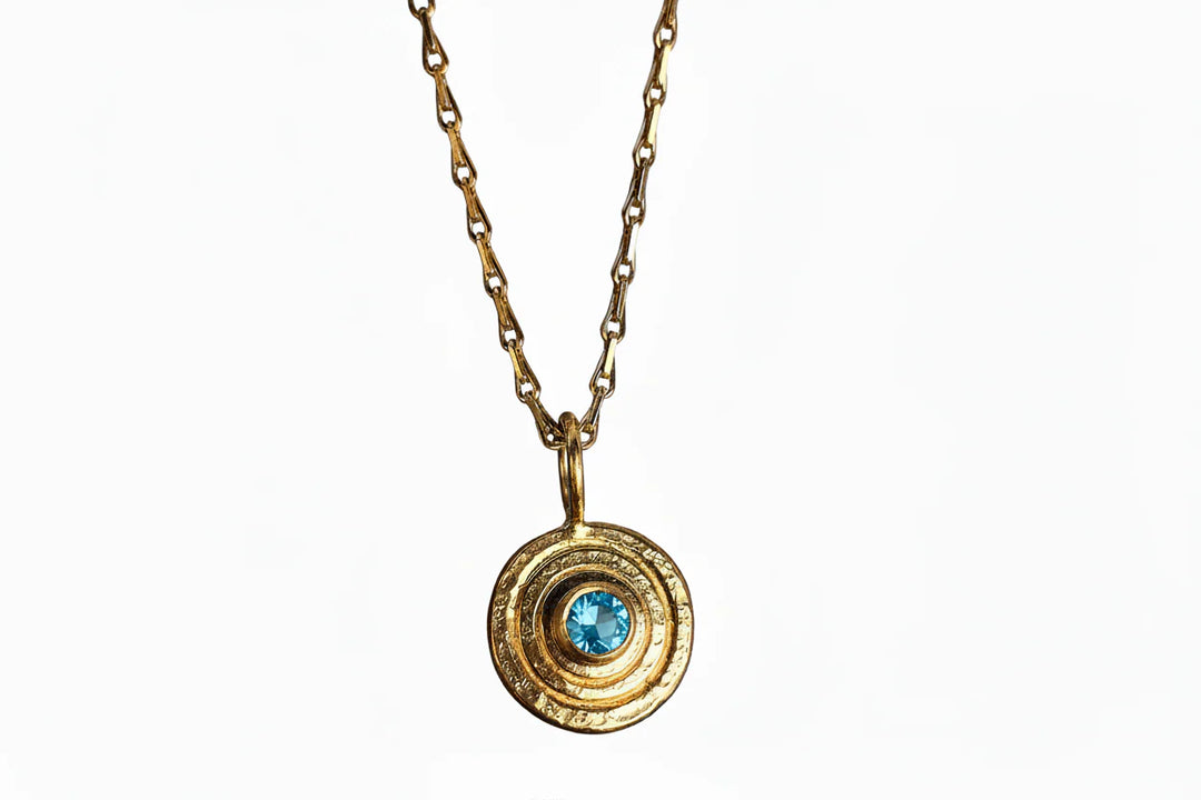 The "Orb Pendant Gold in Blue Topaz" by Caroline Stokesberry-lee features a gold-plated sterling silver chain with a circular pendant of concentric rings and a central blue topaz, shown against a white background.