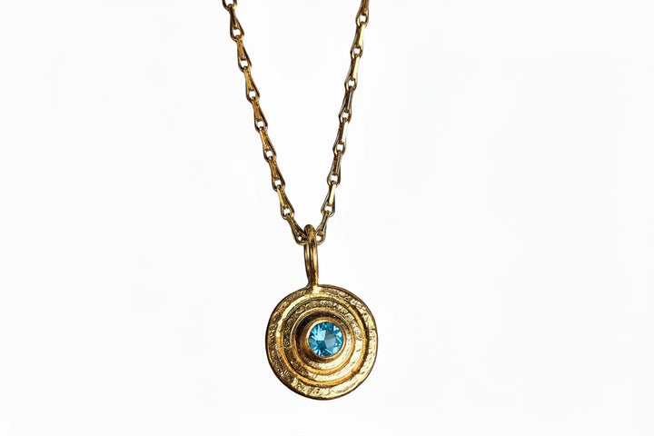 The "Orb Pendant Gold in Blue Topaz" by Caroline Stokesberry-lee features a gold-plated sterling silver chain with a circular pendant of concentric rings and a central blue topaz, shown against a white background.
