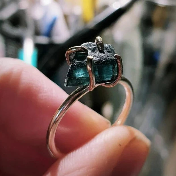 A hand displays the Caroline Stokesberry-lee Raw Green Apatite Gold Filled Ring, featuring a minimalist open prong setting and an uncut green apatite gemstone, with a blurred background for emphasis.