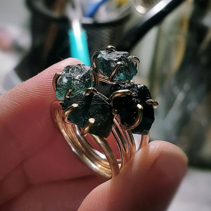 A hand displays three Caroline Stokesberry-lee Raw Green Apatite Gold Filled Rings, each featuring a large, raw green apatite gemstone in a prong setting against a blurred background.