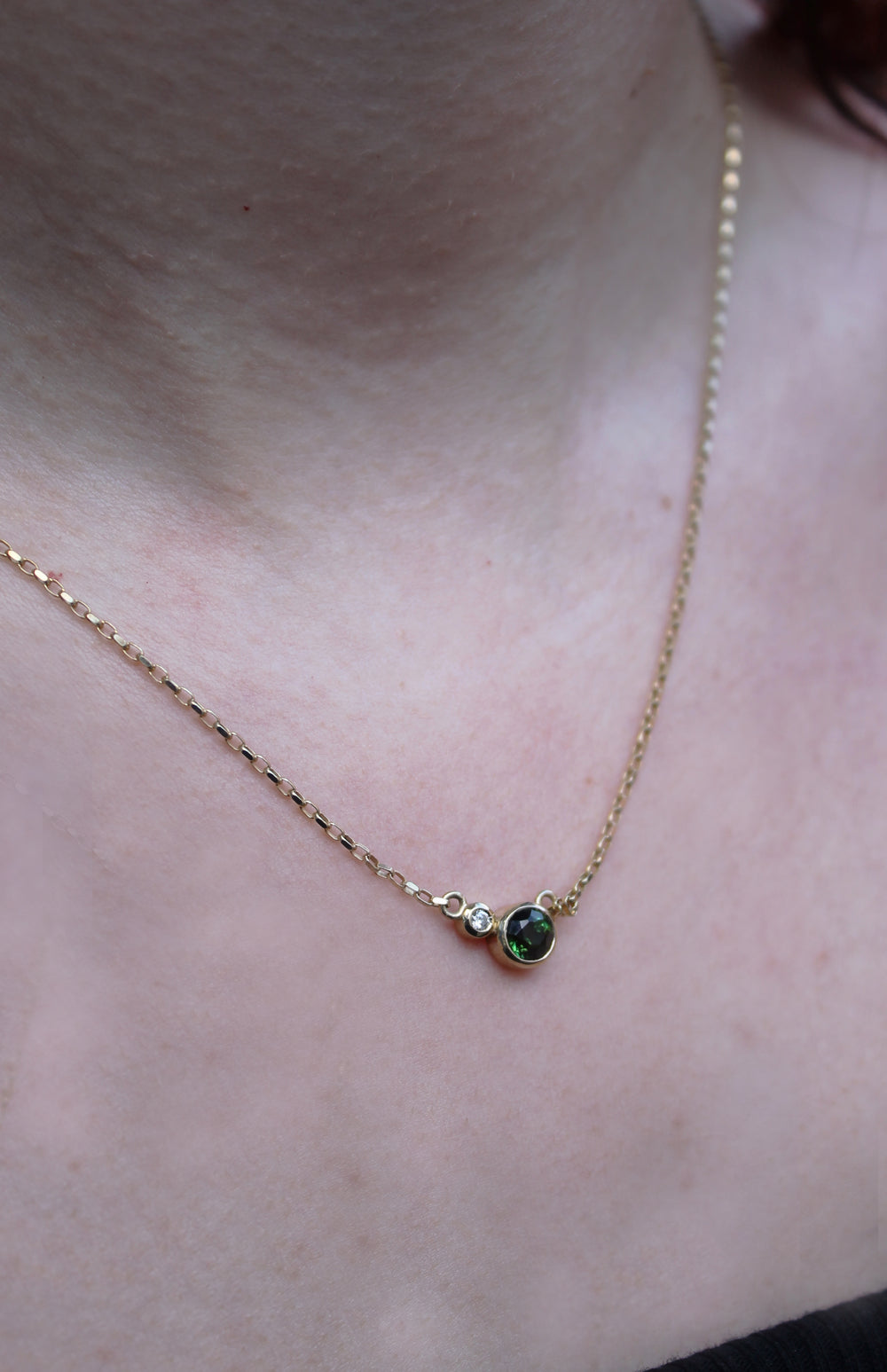 A close-up shows the Madeleine Verdant Embrace 9ct Yellow Gold Necklace, featuring a small circular green tourmaline and lab grown diamond pendant, worn around a neck above a dark top with blurred skin in the background.