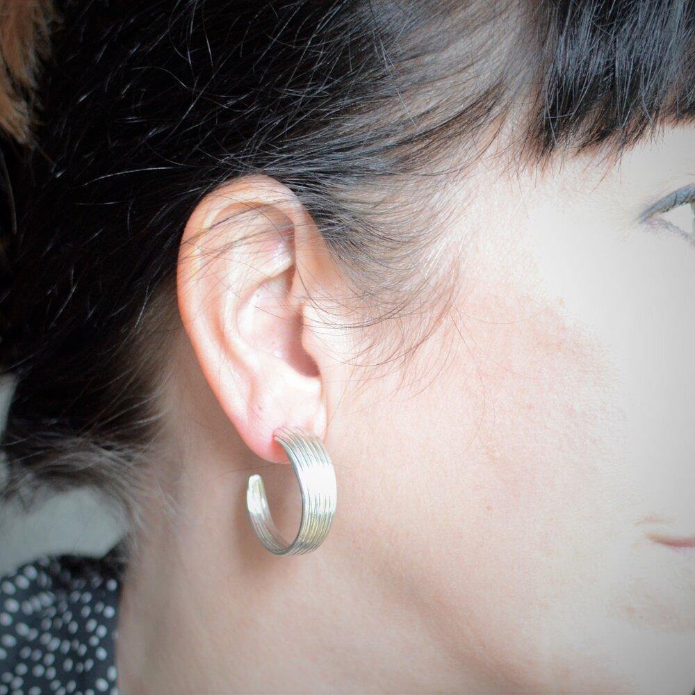 Close-up of a person wearing an Abbie Dixon Mitsuro Hoop Earring with ridged texture in their left ear. The person has dark hair and light skin. The background is blurred, focusing attention on the sterling silver earring and ear.