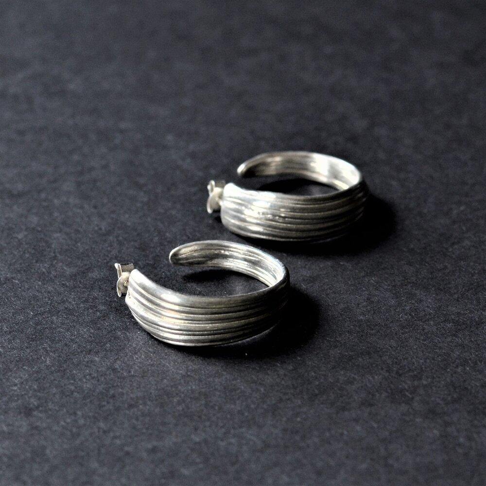 A pair of Mitsuro Hoop Earrings by Abbie Dixon with a ribbed design sit on a dark surface. The earrings are open hoops with a wider band, giving them a bold, textured look. The surface they're on enhances the earrings' metallic shine.