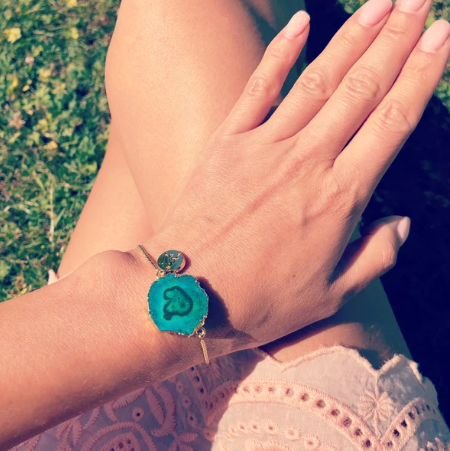 A person with light skin wearing a pink, eyelet-patterned dress is sitting on grass, showing their left hand and wrist adorned with an Aria V Holly Bracelet with a small heart-shaped design in the center. The bracelet also features a small circular charm.