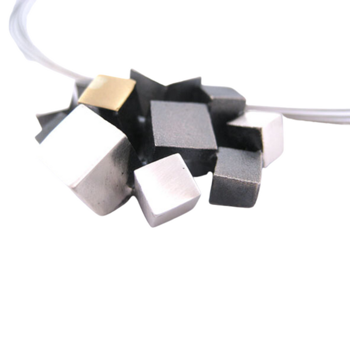 A close-up photo of the Iceland Pendant from MarCucurella, featuring variously sized metallic cubes in shades of silver, black, and gold, clustered together and attached to a thin, transparent wire. The design is minimalist with an industrial aesthetic.