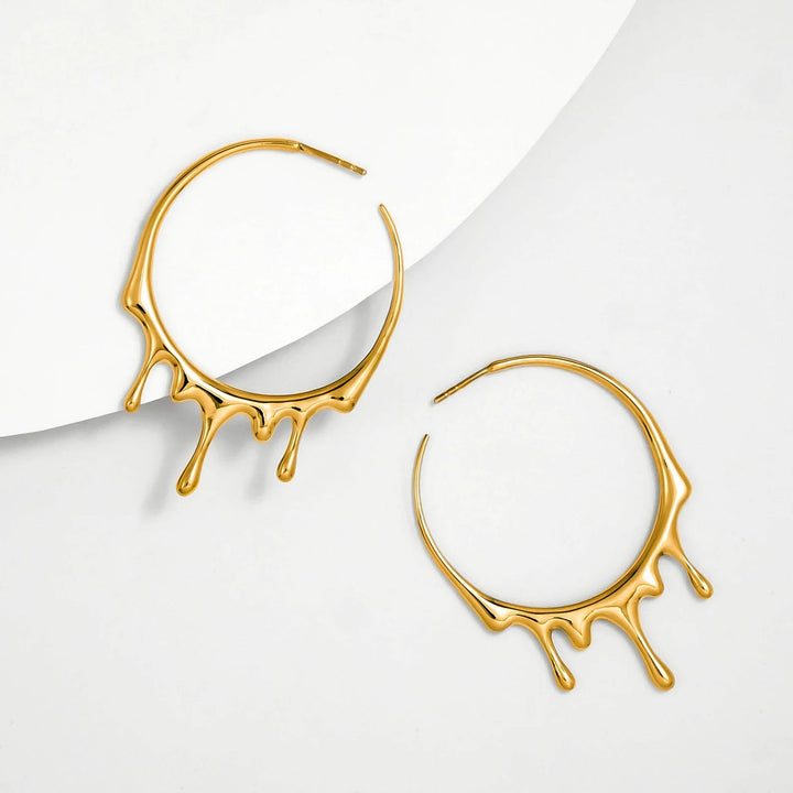 A pair of 18K yellow gold hoop earrings, known as Gold Dripping Circular M Hoop Earrings by Marie June Jewellery, with a unique, artistic design resembling dripping liquid, placed on a white background. The earrings are arranged diagonally, with one earring on the top left and the other on the bottom right.