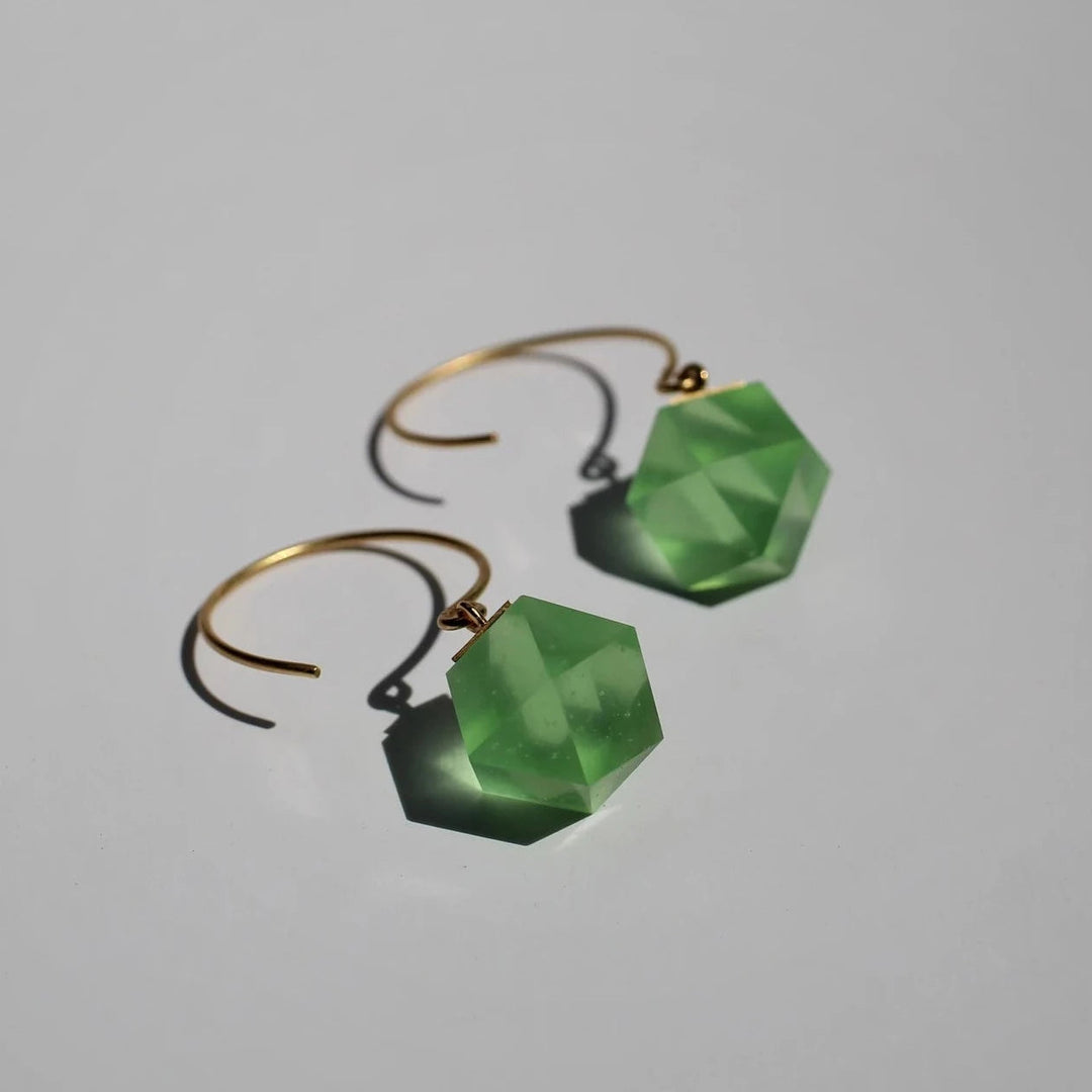 Introducing the GEO Round Hook Earrings from Beira by Irene Reyes: stunning gold hoops adorned with geometric, faceted Bullseye glass in translucent green, shaped like hexagonal prisms casting soft shadows on a light gray background.