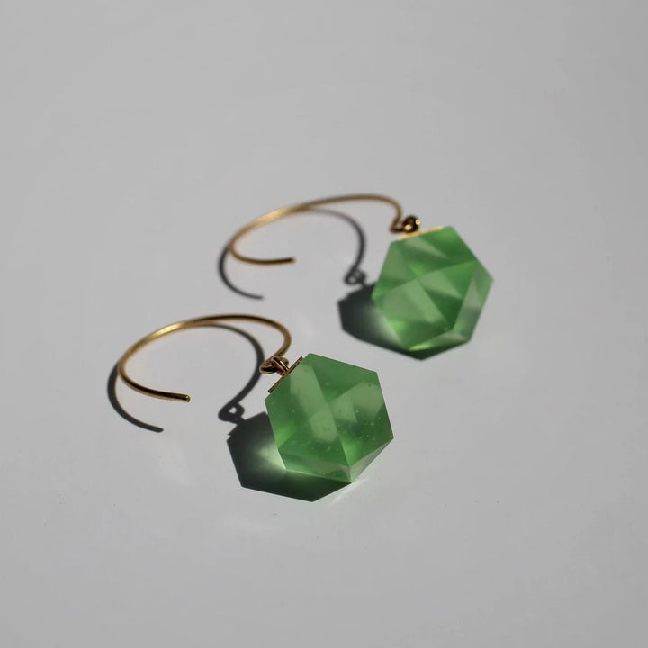 Introducing the GEO Round Hook Earrings from Beira by Irene Reyes: stunning gold hoops adorned with geometric, faceted Bullseye glass in translucent green, shaped like hexagonal prisms casting soft shadows on a light gray background.