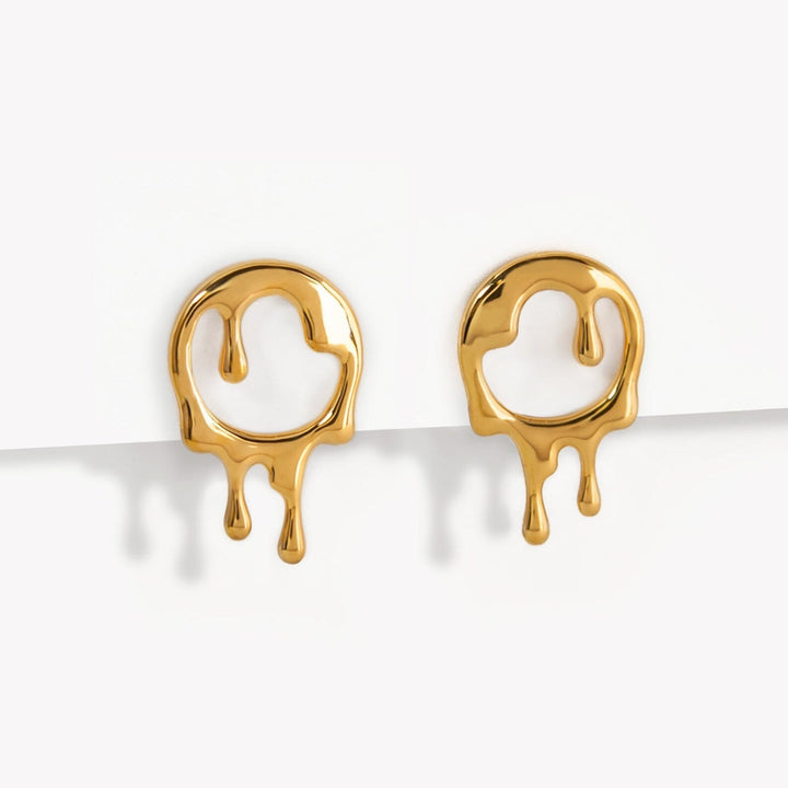 A pair of Dripping Rivulets Stud Earrings Gold by Marie June Jewellery shaped like abstract, dripping smiley faces. The faces feature circular structures with melting effects, extending down into multiple drip forms. The earrings have a polished, shiny surface and are displayed against a white background.