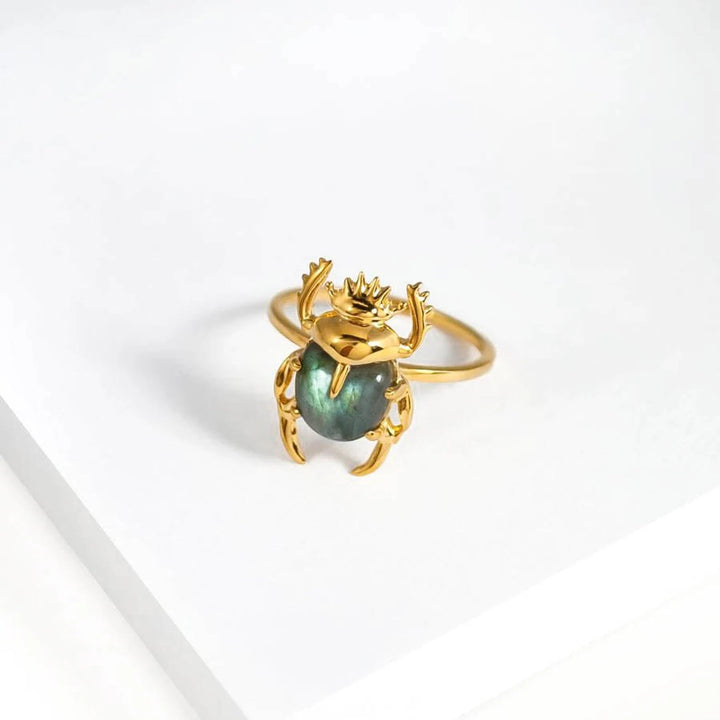 A gold ring featuring a beetle design with an iridescent green gemstone body is displayed on a white surface. The Marie June Jewellery Sacred Scarab Gemstone Ring's head and legs are stylized, and the band's 24k Gold Vermeil finish is plain yet elegant. The overall aesthetic is both whimsical and sophisticated.