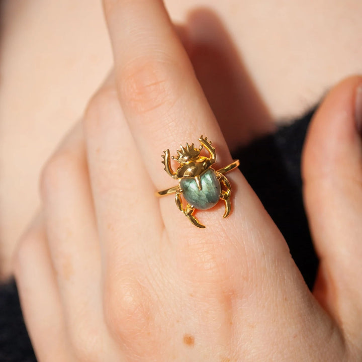 Close-up of a hand wearing a unique Sacred Scarab Gemstone Ring by Marie June Jewellery in 24k Gold Vermeil. The scarab beetle's body is made from a light green, natural hand-carved labradorite. The hand is pointing upward, with the finger wearing the ring prominently displayed. The background is blurred, emphasizing the ring's details.