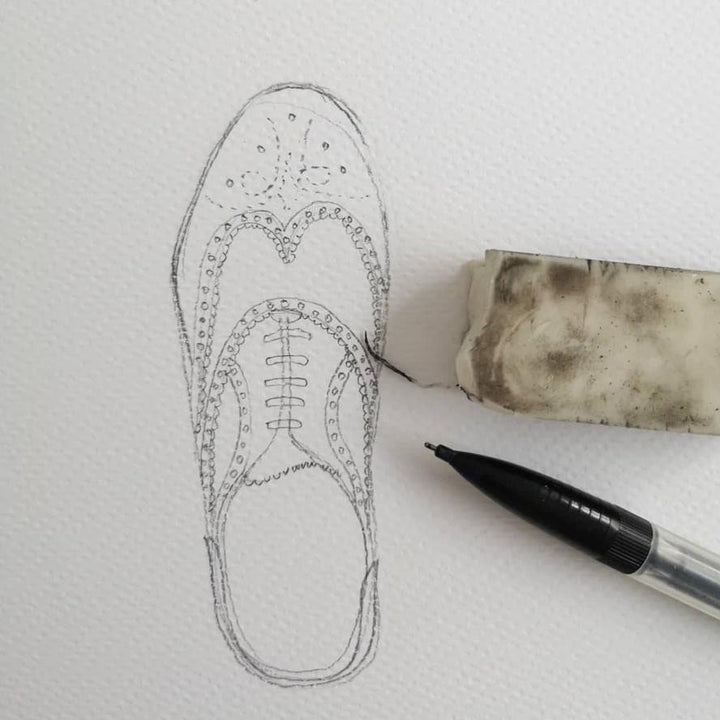 A detailed pencil sketch of an Oxford-style shoe on white paper, with an eraser partially smudged on the right side and a black pen resting beside it. The original illustration features decorative details and laces, perfect for pairing with a beautifully textured card or placed inside a kraft envelope. The product, "KEEP ACTING YOUR SHOE SIZE" by Pickled Pompom, brings this exquisite artwork to life.