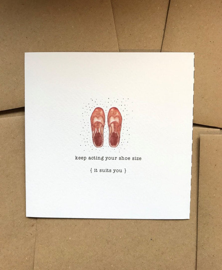 A beautifully textured card shown against a brown background features an original illustration of brown shoes at the top center. Below, text reads: "keep acting your shoe size { it suits you }." It comes tucked in a Kraft envelope. The card is called KEEP ACTING YOUR SHOE SIZE, and it's from the brand Pickled Pompom.