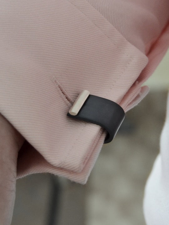 Close-up of a person wearing a light pink dress shirt with Affinor Wrap Around Cufflinks by Filip Vanas securing the cuff. The attire is formal and neatly pressed. The background appears blurred and indistinct, likely an indoor setting.