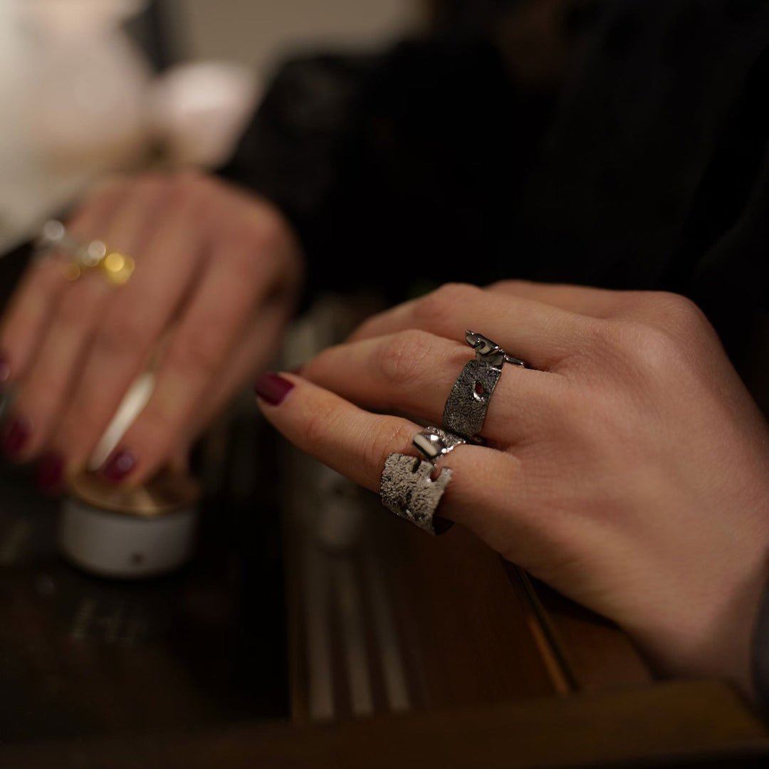 Close-up of two hands with well-manicured nails, one with red nail polish, and three handcrafted silver rings from Theodora D. The hands are resting on a surface, one hand touching a small cylindrical object. The background is blurred, emphasizing the focus on the hands and the Rythmos Black Rhodium Ring by Theodora D.