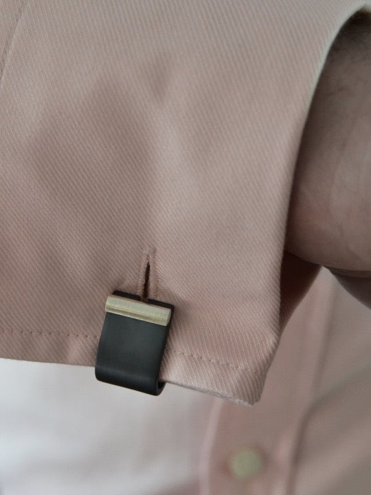 Close-up of a wrist wearing a light pink dress shirt with an innovative cuff link featuring dark gray and silver sterling silver anodized aluminum. The cufflink is attached to the subtly textured diagonal fabric, highlighting part of the elegant Affinor Wrap Around Cufflinks collection by Filip Vanas on the visible cuff and sleeve.