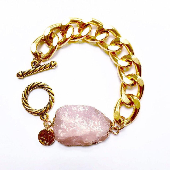 The Cruise Chunky Bracelet from Aria V features large, intertwined gold links and a rose quartz stone, handmade in Dublin. Attached is an irregularly shaped, pale pink stone, and a gold toggle clasp with a twisted design. A small round gold charm with an engraved pattern hangs near the clasp. The background is white.