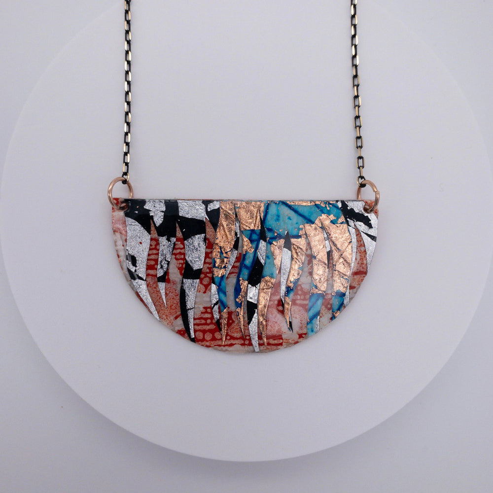 The Bláth Batik Textile Necklace by Rothlú is a handmade piece featuring a half-circle design adorned with an abstract pattern in orange, copper, silver, blue, and black. The pendant, finished with eco resin for a unique texture, hangs from a thin black chain against a plain white background.