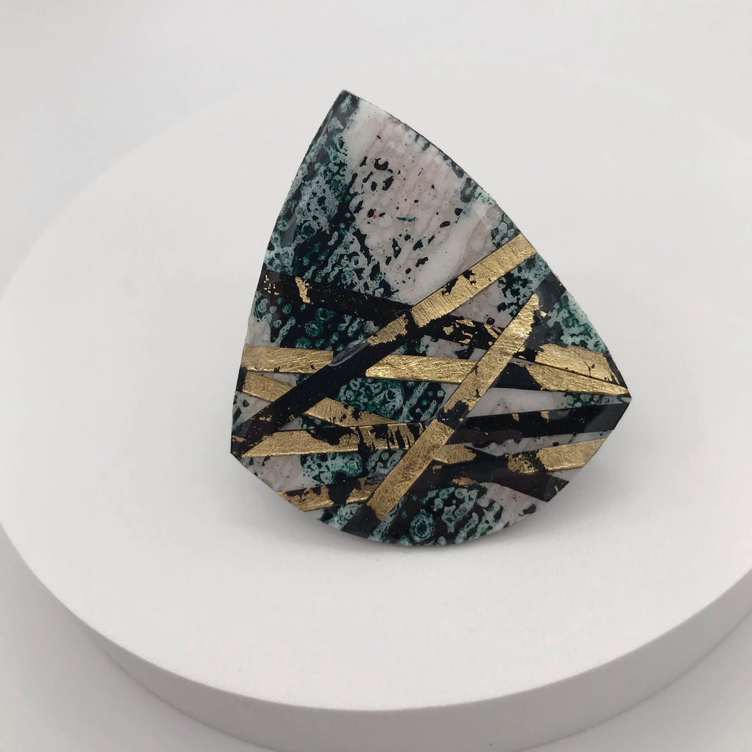 A polished, asymmetrically shaped stone with a mix of green, black, and white hues is displayed on a white circular stand. The stone is adorned with gold diagonal and crisscrossing lines, reminiscent of the batik tjanting technique, giving it a striking and artistic appearance. This is the Crosstown Ring Dark Green/Black/Gold by Rothlú.