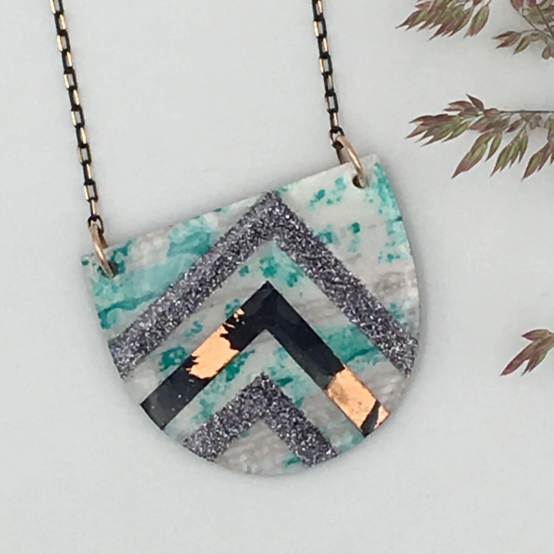 A Rothlú Sayre Batik Textile Necklace in Mint/Black/Copper/Grey Shimmer with a semi-circular pendant featuring a geometric design. The pendant has layers of teal, grey, black, and copper colors, with textures ranging from smooth to glittery. It hangs from a delicate black-gold faceted chain. Some foliage is visible in the background.