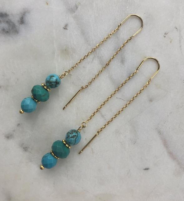 Long gold threader earrings are displayed on a marble surface. Each earring features a delicate 16k gold plated chain with three handcrafted turquoise stone beads in varying shades of blue and green, faceted for a sparkling effect, connected to the end. These are the **Teia Earrings** by **Aria V**.