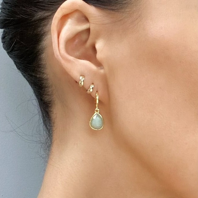 Close-up of a person's ear adorned with elegant jewelry. The person wears two small hoop earrings on the upper part of the ear and a larger drop earring featuring Aria V Amazonite Drop Earrings on the lower part. The background is plain and gray.