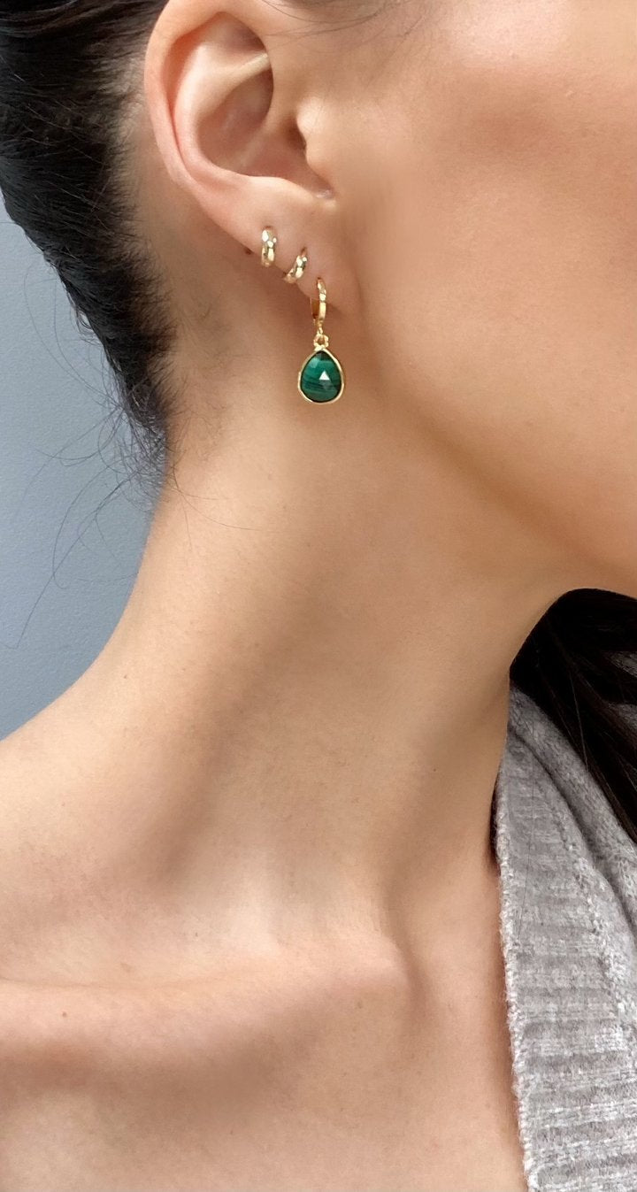 Close-up of a woman's ear adorned with multiple earrings. She is wearing a small 14k gold plated brass hoop earring with a teardrop-shaped Malachite Drop Earrings by Aria V stone, and two smaller, simple gold studs above it. The background is neutral, and a portion of her gray top is visible.