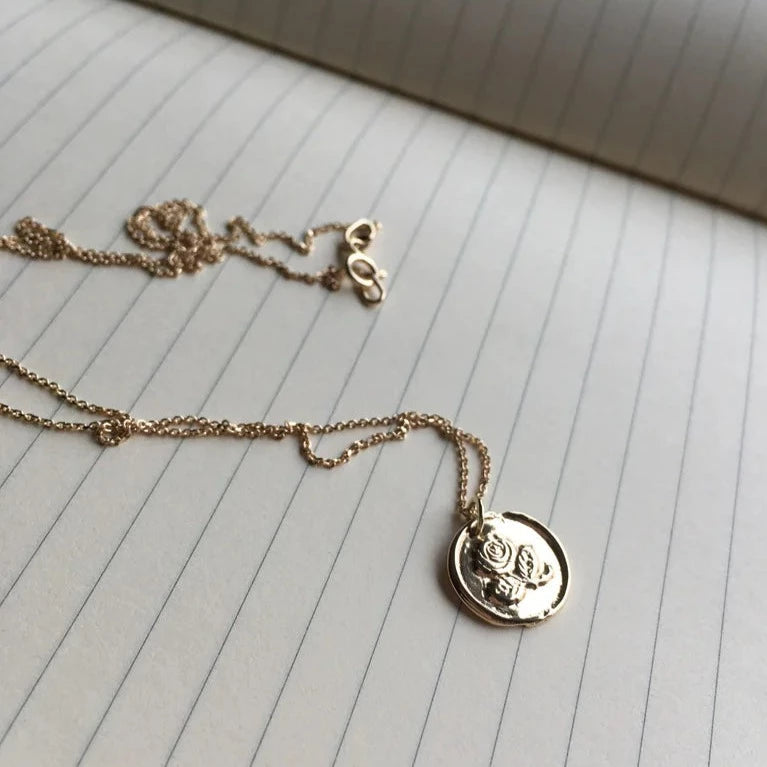 A delicate gold-plated necklace with a circular pendant featuring a raised owl design rests on an open lined notebook. The Irish Rose Necklace Gold from Andrea Mears Jewellery is slightly tangled, and the clasp is visible at the top, adding to the intricate detail of the piece.