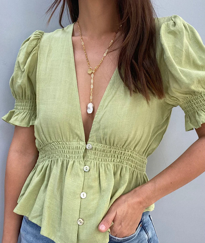 A person wearing a light green blouse with short, puffed sleeves, a deep V-neckline, and buttons down the front. The blouse has a smocked waist and ruffled hem. They are also wearing an Aria V Medea Necklace/Bracelet with a baroque white pearl pendant on a 16k gold electroplated chain and have long, brown hair.