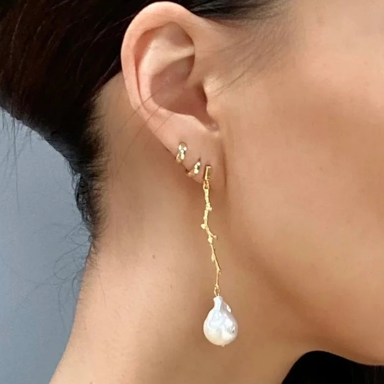 Close-up of a person wearing an earring. The White Medusa Earrings by Aria V feature a gold branch-like design with a large, irregularly shaped white baroque pearl hanging from it. The person has dark hair pulled back and is also wearing multiple small gold studs in the same ear, all handmade in Ireland.