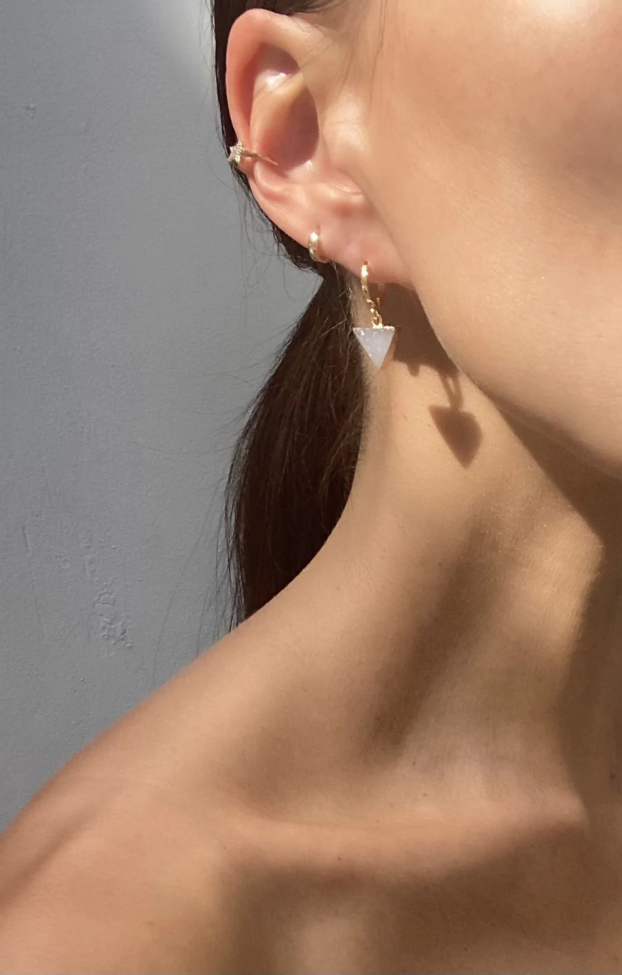 Close-up of a person's ear showcasing two earrings: a 14k gold plated brass hoop at the top and an Aria V Dune Earrings with a white triangular pendant below. The person's hair is tied back, revealing the skin and the earrings clearly against a neutral background.