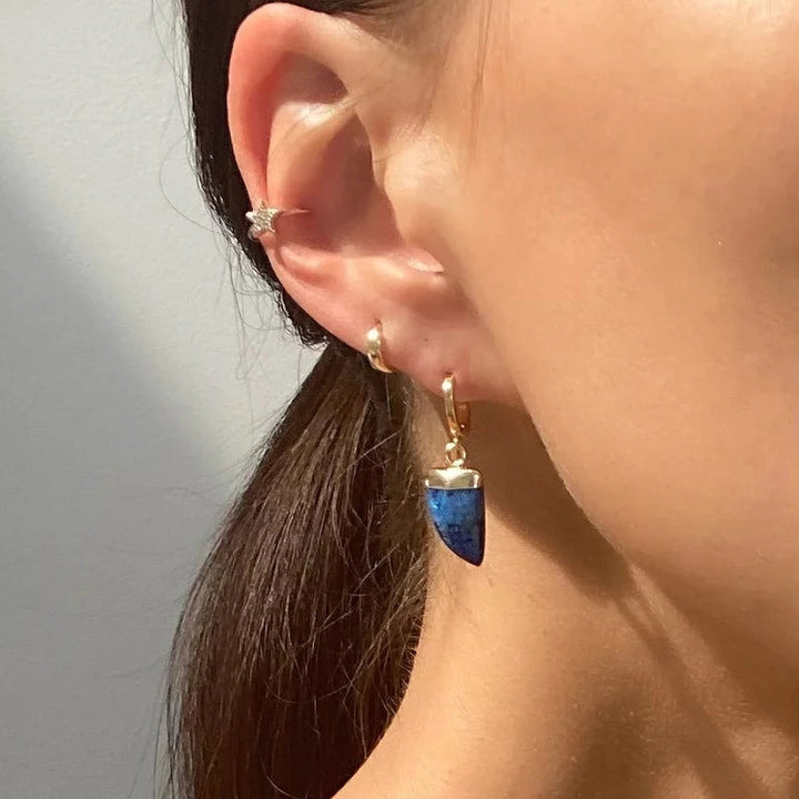 Close-up of a person’s ear wearing multiple Aria V Siren Earrings: a small star-shaped stud near the top, a hoop adorned with Natural Lapis Lazuli and gold pendant, and another small hoop in the earlobe. The person’s dark hair is pulled back, showcasing the earrings clearly against the background.