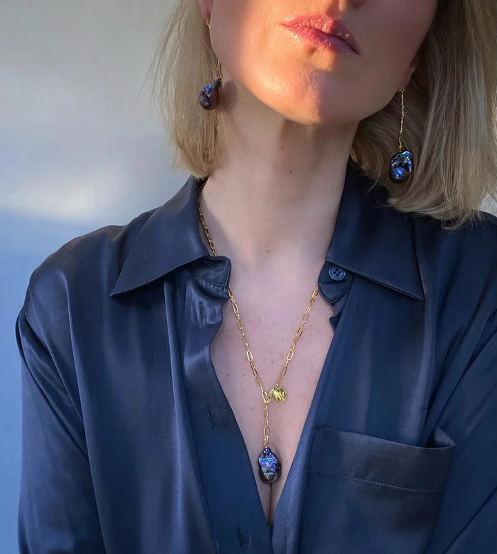 Close-up of a woman wearing a dark blue silk shirt, paired with stunning Petrol Medusa Earrings by Aria V and a 16k gold chain necklace adorned with coordinating gemstone pendants. The lighting casts a warm glow on her face and upper body.