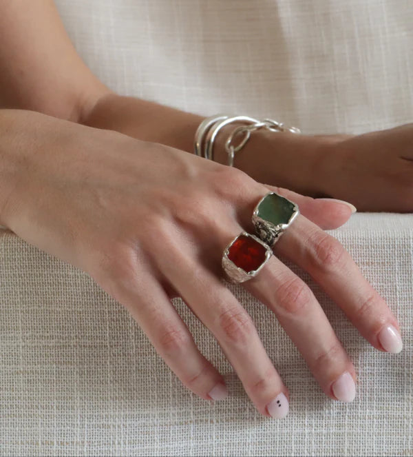 Close-up of a hand on textured fabric adorned with two Essence Silver Rings by Inó Jewellery, featuring semi-precious green and red stones. A silver bracelet is also visible against the cream background.