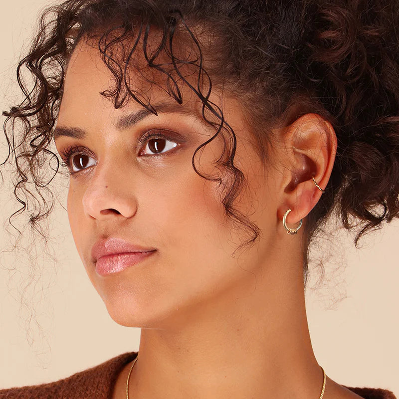 A woman with curly hair, wearing Scribble & Stone’s Infinity Midi Hoop Earrings and a brown top, gazes to the side with a neutral expression against a light beige background.