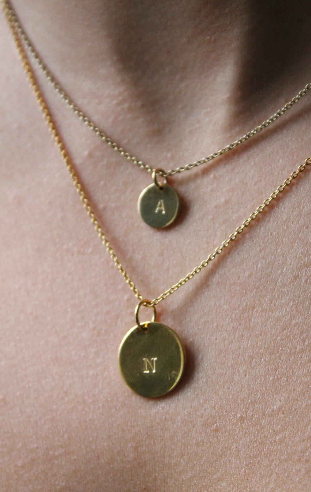 Close-up of two silver necklaces on a person’s neck, each with a round hand-stamped charm from Personalised Charms; the upper pendant is engraved with "A" and the lower, larger one is the Personalised Large Disc Charm in Silver engraved with "N".