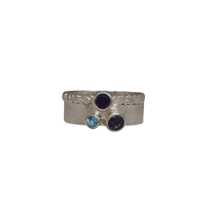 A frosted silver ring with a wide band featuring three faceted stones on top. The central gem is a deep blue color, flanked by two smaller purple stones on either side. The ring has a textured design around the band. This stunning piece is the Iolite, Amethyst & Aqua Marine Wing Ring by Leoma Drew.