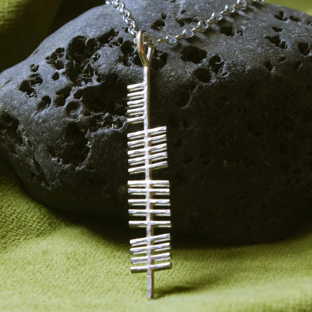 An Ireland (Erin) Pendant inscribed with the Ogham alphabet from Ogham Treasure hangs on a chain against a background of black porous rock and green cloth. The linear design of the Ogham letters adds an element of ancient Irish script to this elegant piece of jewelry.