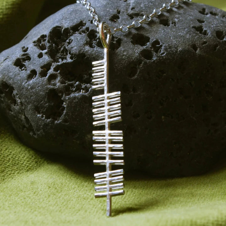 An Ireland (Erin) Pendant inscribed with the Ogham alphabet from Ogham Treasure hangs on a chain against a background of black porous rock and green cloth. The linear design of the Ogham letters adds an element of ancient Irish script to this elegant piece of jewelry.