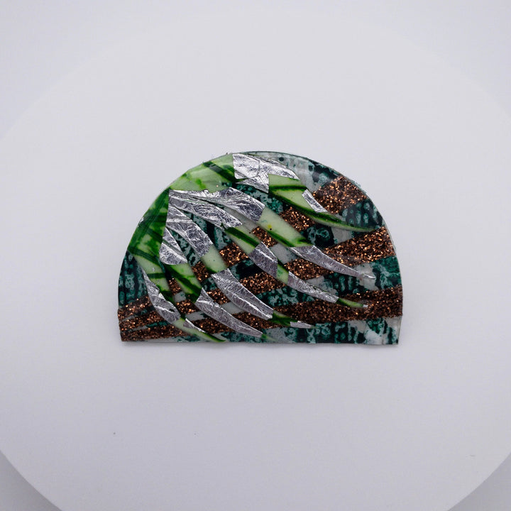 The Bláth Batik Textile Brooch by Rothlú is an abstract, half-circle art piece showcased on a white background. It features overlapping layers enhanced with an eco resin finish in metallic silver, glittery brown, green, and multicolored textures that create a visually dynamic pattern.