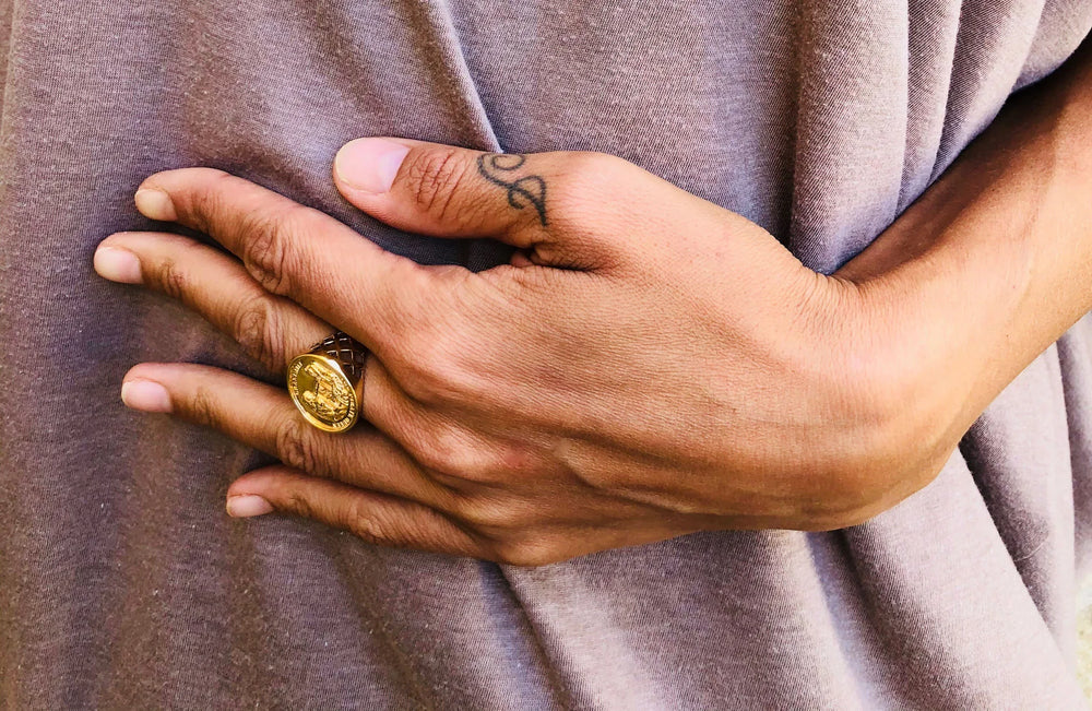 A close-up of a left hand with a small finger tattoo rests on light brown fabric, wearing the handmade Jennifer Kinnear Grace O’Malley Sovereign Ring with an engraved design.