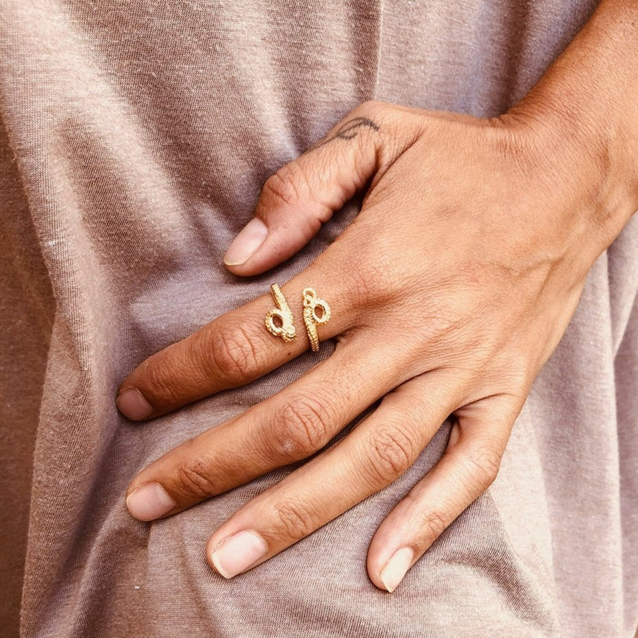 A person's hand with a visible tattoo on the index finger rests on a beige fabric. They are wearing two gold-colored rings shaped like snakes, one on the index and one on the middle finger, both featuring intricate detailing. On the ring finger, an adjustable solid silver Octopus Ring by Jennifer Kinnear adds a unique touch.