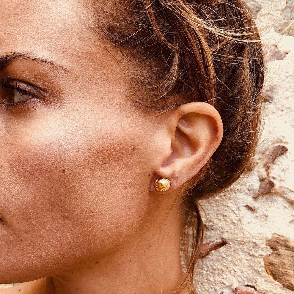 Close-up of the right side of a woman's face, showing her ear adorned with a Jennifer Kinnear Tiny Cockle Shell Stud Earring. The woman has light brown hair tied back, and the background consists of a textured stone wall.