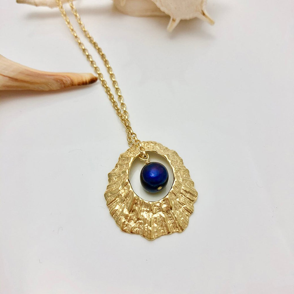 The Hollow Limpet Shell Necklace with Pearl Large by Jennifer Kinnear features a textured, oval-shaped gold frame and a striking dark blue spherical stone at the center. This eye-catching necklace boasts a delicate sterling silver chain and is showcased on a white surface adorned with small seashell decorations nearby.