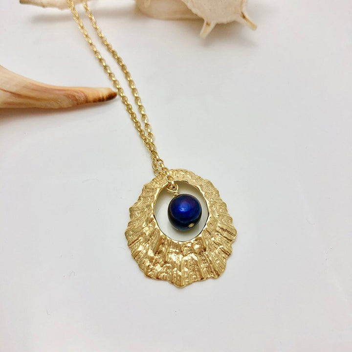 The Hollow Limpet Shell Necklace with Pearl Large by Jennifer Kinnear features a textured, oval-shaped gold frame and a striking dark blue spherical stone at the center. This eye-catching necklace boasts a delicate sterling silver chain and is showcased on a white surface adorned with small seashell decorations nearby.